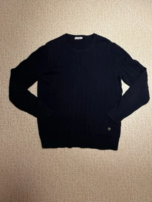 Mörkblå stickad tröja J.Lindeberg - Snygg mörkblå stickad tröja från J.Lindebergmed diskret strukturmönster och rund hals. Tröjan har långa ärmar, ribbade muddar och en liten logodetalj vid nederkanten. Perfekt till jeans eller chinos. Storleken är XL men av någon andledning sitter precis som M. Priset kan diskuteras. Hör av dig vid absolut minsta fråga!