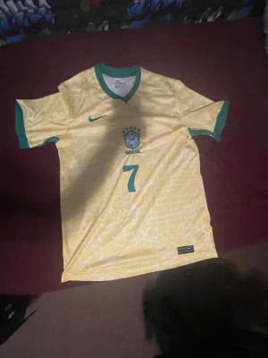 Brasilien Nike fotbollströja gul - Gul Brasilien fotbollströja från Nike med gröna detaljer vid krage och ärmslut. Nummer 7 på framsidan och Brasilien-emblem på bröstet. Tillverkad i lätt och ventilerande material, perfekt för fotbollsträning eller match.