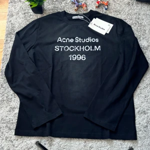 Svart långärmad topp Acne Studios - Svart långärmad topp från Acne Studios med tryck 'Acne Studios STOCKHOLM 1996' i vitt framtill. Klassisk rund halsringning och rak passform. Perfekt för dig som gillar stilrena och tidlösa plagg.