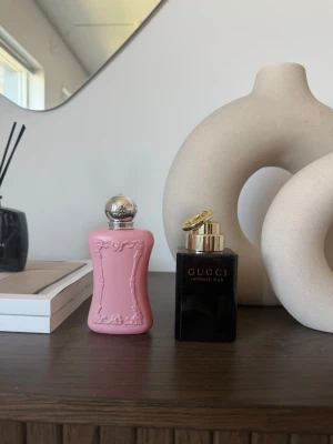 Gucci Intense Oud parfym & rosa flaska - Två parfymer: Gucci Intense Oud i en elegant svart och guldig rektangulär flaska samt en rosa parfymflaska med silverfärgad rund kork och dekorativa detaljer. Båda flaskorna har lyxig design och passar perfekt för dig som gillar exklusiva dofter.