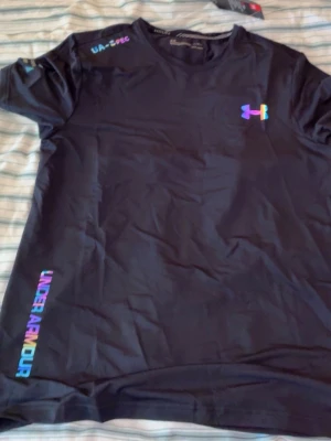 Under Armour t-shirt med regnbågslogga - Du får båda för 600.Två snygga Under Armour t-shirts i svart och vitt med regnbågsfärgad logga och text. Klassisk passform, korta ärmar och rund hals. Perfekt för dig som vill ha en sportig och modern look. Materialet är mjukt och andas, troligtvis polyester.