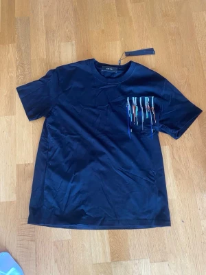 Svart AMIRI t-shirt med färgstänk - Snygg svart t-shirt från AMIRI med coolt tryck framtill där loggan har färgstänk i blått, orange, grönt och vitt. Klassisk passform och rund halsringning. Perfekt för dig som vill sticka ut med en trendig streetstyle-look.