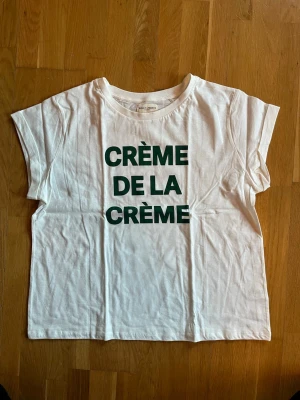 Vit t-shirt Holly & Whyte XL - Vit t-shirt från Holly & Whyte by Lindex i storlek XL. Tryck i grönt med texten 'CRÈME DE LA CRÈME' på framsidan. Klassisk passform med korta ärmar och rund hals. Tillverkad i mjuk bomull, perfekt för en avslappnad look.