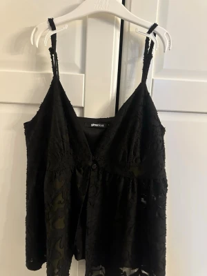 Svart spetslinnetopp från Gina Tricot - Snygg svart linnetopp från Gina Tricot med tunna axelband och v-ringning. Toppen är dekorerad med spetsdetaljer och har en knapp framtill. Perfekt för dig som gillar en feminin och trendig look. Aldrig använd!