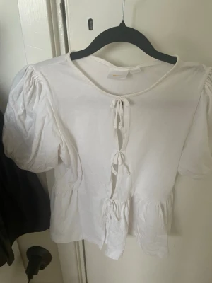 Vit blus med knyt från Gina Tricot - Supersöt vit blus från Gina Tricot med puffärmar och knyt framtill. Blusen har en lätt volang nertill och är gjord i mjuk bomull. Perfekt för dig som gillar en romantisk och trendig stil.
