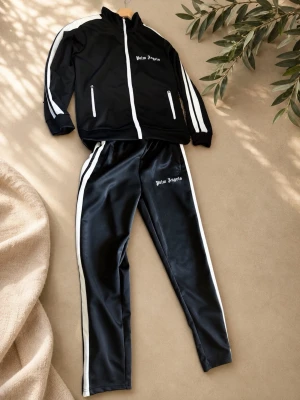 Palm Angels svart tracksuit set - Svart tracksuit från Palm Angels med vita ränder längs ärmar och ben. Jackan har dragkedja, ståkrage och två fickor med dragkedja. Byxorna har resår i midjan och vita sidoränder. Båda delarna har Palm Angels-logga tryckt i vitt.