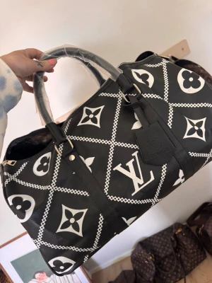 Weekendbag  - Weekendbag i svart  med vita monogram och grafiska detaljer. Väskan har dubbla handtag och dragkedja upptill. Perfekt för dig som vill sticka ut med en trendig och lyxig accessoar.