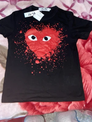 Svart t-shirt PLAY med rött hjärta - Svart t-shirt från PLAY med ett stort rött hjärta och ögon på bröstet, omgiven av färgstänk. Klassisk passform och rund hals. Perfekt för dig som vill sticka ut med en cool och lekfull design.