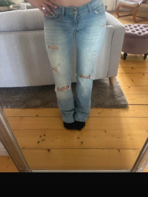 Ljusblå slitna bootcut jeans - Säljer ett par ljusblå bootcut jeans med flera slitningar och hål på benen. Jeansen har klassisk femficksdesign och låg midja. Perfekta för en avslappnad och trendig look.