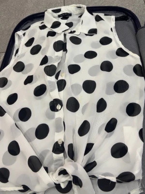 Vit ärmlös blus med svarta prickar - Snygg och luftig ärmlös blus från Zara i vit transparent chiffong med stora svarta prickar. Blusen har klassisk krage, knäppning framtill och knytning nedtill för en trendig look. Perfekt till jeans eller kjol för en cool stil.