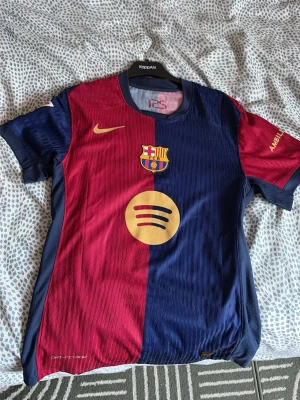 FC Barcelona matchtröja Lamine Yamal - Säljer en officiell FC Barcelona matchtröja från Nike i röd och marinblå med klubbmärke, Spotify-logga och Lamine Yamal 19 på ryggen. Tröjan har UNHCR-tryck och är tillverkad i lätt, ventilerande material perfekt för fotbollsträning.