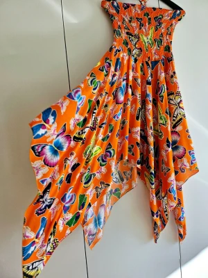 Orange klänning med fjärilsmönster - Färgstark orange klänning med smock upptill och asymmetrisk nederkant. Klänningen har ett iögonfallande fjärilsmönster i flera färger och är ärmlös. Perfekt för dig som vill sticka ut med en unik och somrig look. - Köparen betalar för frakten 