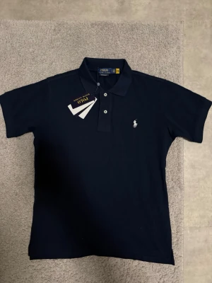 Marinblå pikétröja från Polo Ralph Lauren - Klassisk marinblå pikétröja från Polo Ralph Lauren med vit broderad logga på bröstet. Tröjan har krage, knappar framtill och korta ärmar. Perfekt till jeans eller shorts för en clean och stilren look.