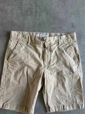Beige shorts från GANT HENLEY - Klassiska beige shorts från GANT HENLEY med normal passform. De har fickor fram och bak, bälteshällor och stängs med knapp och dragkedja. Perfekta för varma dagar och enkla att matcha med olika stilar.