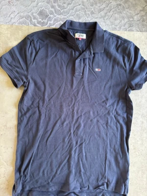 Mörkblå pikétröja från Tommy Hilfiger - Klassisk mörkblå pikétröja från Tommy Hilfiger med liten logga på bröstet och krage med knappar. Perfekt till jeans eller shorts för en avslappnad men snygg stil.