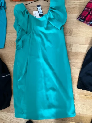 Turkos klänning med volang från Vila - Snygg turkos klänning från Vila med volangdetaljer upptill. Klänningen är ärmlös och har en rak, enkel passform som ger en stilren look. Perfekt för dig som vill sticka ut med färg och elegans.