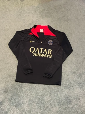 PSG träningströja från Nike - Snygg svart långärmad träningströja från Nike med PSG-logga på bröstet och Qatar Airways-tryck i gult. Tröjan har halv dragkedja och röda detaljer vid kragen. Perfekt för fotbollsträning eller sportiga dagar.