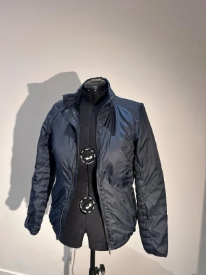 Mörkblå pufferjacka med hög krage - Snygg mörkblå pufferjacka med dragkedja framtill och hög krage. Jackan har en klassisk quiltad design och två sidofickor med dragkedja. Perfekt för kalla dagar och enkel att matcha med olika outfits.