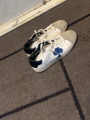 Vit sneaker med blå detalj The Gallerist - Snygga vita sneakers från The Gallerist med blå klöverdetalj på sidan och mörkblå häl. Skorna har klassisk låg profil, vita snören och grå tåparti. Perfekta för dig som gillar unik design och streetstyle.