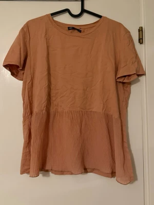Persikofärgad blus från Zara - Säljer en persikofärgad blus från Zara med korta ärmar och rund halsringning. Nedre delen av blusen har plisserade detaljer som ger en snygg och luftig känsla. Perfekt för dig som gillar en avslappnad men trendig stil.