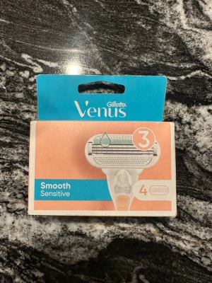 Gillette Venus rakblad Smooth Sensitive - Förpackning med fyra Gillette Venus Smooth Sensitive rakblad. Rakbladen har tre blad och är utformade för känslig hud. Förpackningen är i blått och persikofärgat med tydlig märkning. Passar Venus-rakhyvlar.