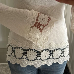 Vit topp med spetsdetaljer - Snygg vit långärmad topp med genombrutna spetsdetaljer och blommönster vid ärmslut och nederkant. Tröjan har utsvängda ärmar och är tillverkad i ett tunt, mjukt material som ger en feminin och trendig look. Helt ny med prislapp kvar!!!