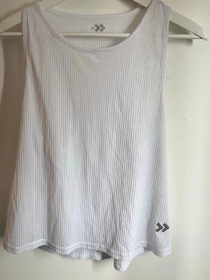 Vitt träningslinne  - Snygg vit linnetopp från Soc med tunna vertikala ränder och sportig design. Toppen är ärmlös med brottarrygg och har en liten logga nertill. Perfekt för träning eller en avslappnad look.