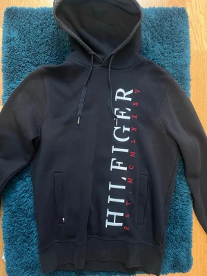 Svart hoodie från Tommy Hilfiger - Snygg svart hoodie från Tommy Hilfiger med stor vertikal logga i vitt och rött på framsidan. Klassisk huva med dragsko och två fickor framtill. Mjuk bomullsblandning som är perfekt för en avslappnad stil. Köpt i Stockholm år 2023 i Tommy hilfigers officiella butik.