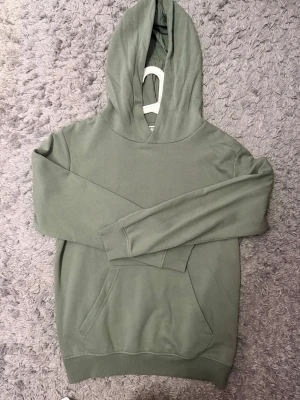 Grön hoodie från Kappahl - Snygg grön hoodie från Kappahl med klassisk huva och stor känguruficka framtill. Mjuk bomullskänsla och ribbade muddar vid ärmslut och nederkant. Perfekt för en chill och avslappnad stil. Passar mig som har XS