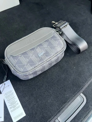Grå axelväska från Lacoste - Snygg grå axelväska från Lacoste med mönstrad textil och detaljer i läderimitation. Väskan har justerbar axelrem, dragkedja och ett diskret Lacoste-logotyp framtill. Perfekt storlek för det viktigaste och stilren design.
