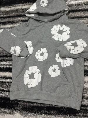 Grå hoodie med vita blommor - Snygg grå hoodie från Denim tears med stora vita blommönster tryckta över hela tröjan. Hoodien har en klassisk huva och känguruficka framtill. Perfekt för en avslappnad och trendig stil.