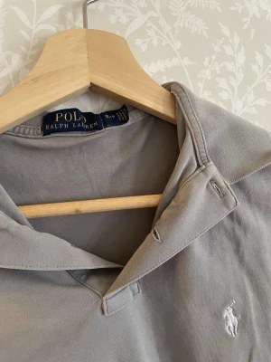 Grå pikétröja från Ralph Lauren - Klassisk grå pikétröja från Ralph Lauren med korta ärmar och vit broderad logga på bröstet. Tröjan har krage och är tillverkad i mjuk bomull, perfekt för en avslappnad och stilren look.