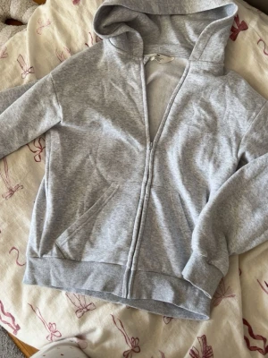Ljusgrå zip-hoodie med huva - En basic ljusgrå hoodie med dragkedja och huva. Tröjan har ribbade muddar vid ärmslut och nederkant samt två fickor framtill. Perfekt för en avslappnad och stilren look. Materialet är mjukt och skönt, troligtvis bomullsblandning.