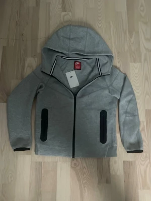 Nike tech grå - Snygg grå hoodie från Nike med hel dragkedja och svarta detaljer. Två fickor med dragkedja framtill och svart Nike-logga på bröstet. Hoodien har huva och svarta muddar vid ärmsluten. Perfekt för en sportig och avslappnad stil.