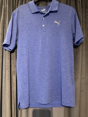 Blå pikétröja från Puma - Snygg blå pikétröja från Puma med klassisk krage och två knappar framtill. Tröjan har korta ärmar och ett diskret Puma-logotyp på bröstet. Perfekt för en sportig och avslappnad stil.