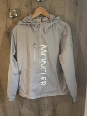 Ljusgrå vindjacka från Moncler - Snygg ljusgrå vindjacka från Moncler med stor logga framtill. Jackan har huva, dragkedja, två sidofickor med dragkedja och en bröstficka. Tillverkad i ett lätt och smidigt material som passar perfekt för blåsiga dagar. Hör av dig innan köp!