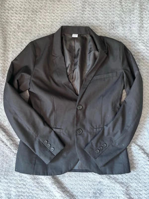 Svart klassisk kavaj med två knappar - Stilren svart kavaj med två knappar framtill och smala slag. Kavajen har tre knappar vid ärmsluten och två fickor framtill. Figursydd modell med helfodrad insida. Perfekt för en snygg och clean look.