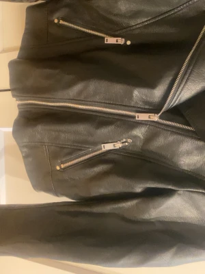 Svart skinnjacka med silvriga dragkedjor - Snygg svart skinnjacka med klassisk bikerstil och silvriga dragkedjor framtill och på fickorna. Jackan har bred krage och detaljerade sömmar som ger en cool look. Perfekt för dig som vill ha en tidlös och edgy stil.