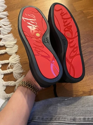 Christian Louboutin sneakers  - Snygga sneakers från Christian Louboutin med den ikoniska röda sulan och svart ovandel. Skorna har snörning och en stilren design som sticker ut med den klassiska signaturen under sulan. Perfekta för dig som vill ha något exklusivt och trendigt.