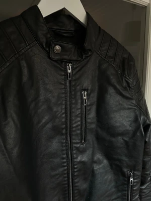 Skinnjacka herr - Svart skinnjacka med ståkrage och dragkedja framtill. Jackan har quiltade detaljer på axlarna, tre fickor med dragkedja och tryckknapp vid hals och nederkant. Konstläder, storlek S💫
