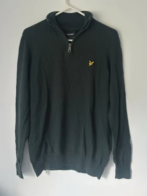 Svart halvzip-tröja från Lyle & Scott - Svart stickad tröja från Lyle & Scott med halv dragkedja och gul logga på bröstet. Tröjan har ribbade muddar vid ärmslut och nederkant samt hög krage. Perfekt för lager-på-lager och snygg till jeans.