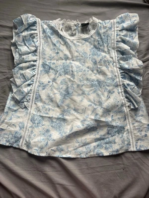 Blommig volangblus från Gina Tricot - Supersöt blus från Gina Tricot med blått blommönster på vit botten. Blusen har volanger längs ärmhålen, spetsdetaljer vid halsen och en rak passform. Materialet känns lätt och luftigt, perfekt för en fräsch look.