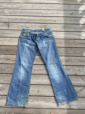 Blå bootcut jeans - Säljer ett par blå bootcut (sitter dock mer baggy) jeans från med snygga slitningar och broderad bakficka. Jeansen har klassisk femficksdesign, låg midja och är tillverkade i denim med lätt stretch. Perfekta för dig som gillar en avslappnad och trendig look.