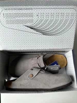 Grå tofflor från Birkenstock - Snygga grå tofflor från Birkenstock i mjuk mocka med justerbart spänne och klassisk korksula. Perfekta för chill dagar hemma eller när du vill ha något bekvämt på fötterna. Ikonisk Birkenstock-logga på sidan och skön innersula.