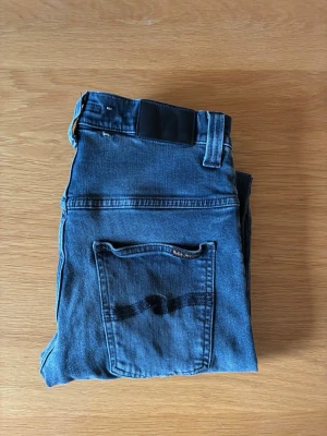 Svarta jeans från nudie - Säljer ett par klassiska svarta jeans från nudie med raka ben och normal midja. Jeansen har fem fickor och en svart patch bak i midjan. Materialet är slitstark denim i bomull och bakfickan har en diskret nudie logga.