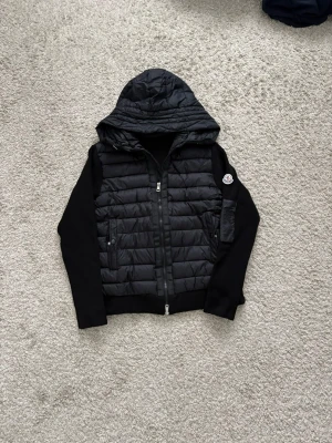 Svart cardigan från Moncler - Svart pufferjacka från Moncler med quiltad kropp och mjuka, långärmade ärmar. Jackan har en stor huva, två fickor med knapp och dragkedja framtill. Moncler-logga på ärmen ger en exklusiv vibe. Perfekt för kalla dagar.