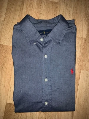 Mörkblå skjorta från Ralph Lauren - Klassisk mörkblå skjorta från Ralph Lauren med knappstängning och röd broderad logga på bröstet. Skjortan har lång ärm och normal passform, tillverkad i mjuk bomull. Perfekt för en stilren look.