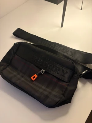 Burberry svart axelväska med logga Helt ny - Snygg svart axelväska från Burberry med klassiskt rutmönster i mörka toner och bred axelrem med präglad logga. Väskan har en orange dragkedjedetalj och är tillverkad i slitstarkt tygmaterial. Perfekt för att bära det viktigaste med stil. 