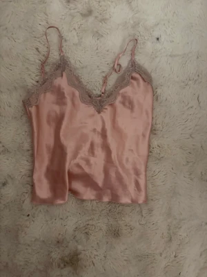 Rosa satinset med spetsdetaljer - Säljer ett sött tvådelat set i ljusrosa satin med tunna axelband och spetskant på linnet samt matchande shorts med spets längs bensluten. Perfekt för en mysig kväll hemma. Materialet är glansigt och mjukt mot huden.