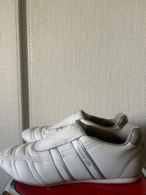 Vita sneakers från Slazenger - Stilrena vita sneakers från Slazenger med silverdetaljer och diskreta ränder på sidorna. Skorna har en låg profil och är tillverkade i syntetmaterial med quiltade sömmar framtill. Perfekta för en clean och sportig look.